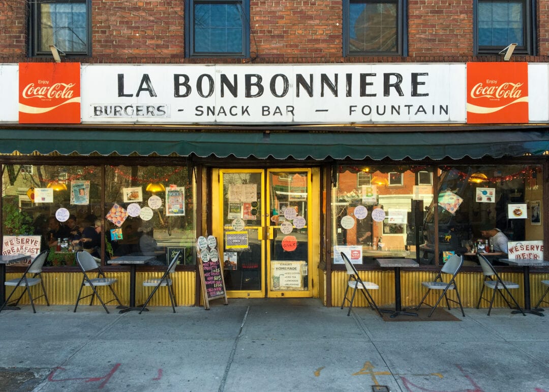 the outside of a diner with a black and white hand painted sign that says "la bonbonniere" flanked by two red coca cola signs