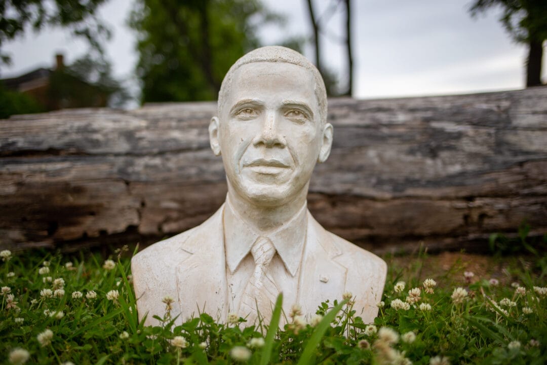 a small white bust of Barack Obama sits in the grass in front of a log