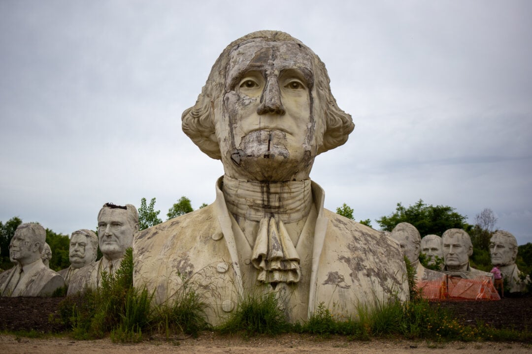 a large bust of george washington crumbling in a field in front of several other similar busts of u.s. presidents