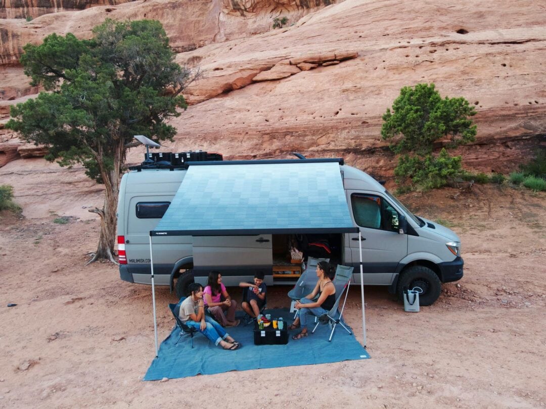 A van-style RV with an awning extended to offer shade to people on the ground.